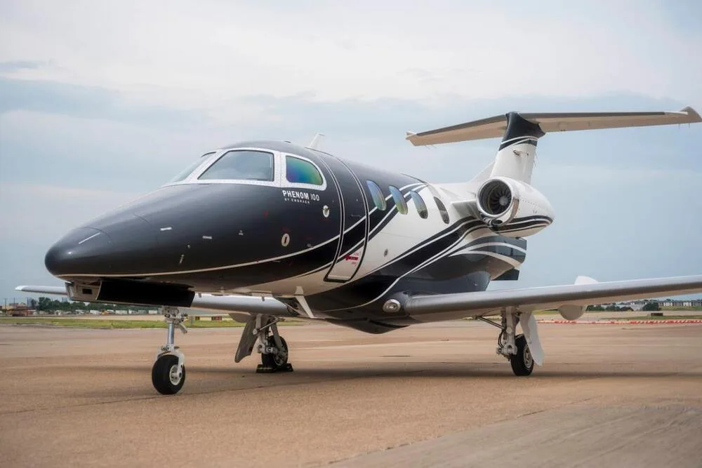 A front-side view of a black and white Embraer Phenom 100 business jet parked on an airport tarmac.