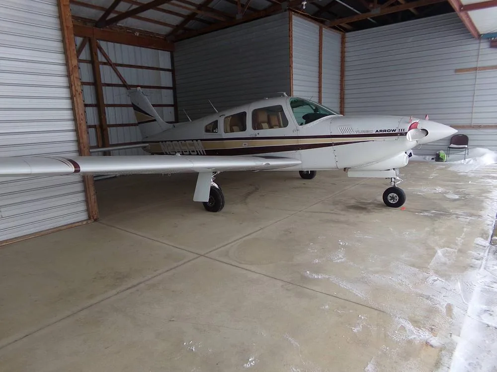 A white Piper Turbo Arrow III aircraft with brown and gold stripes is parked inside a hangar with some snow visible outside.