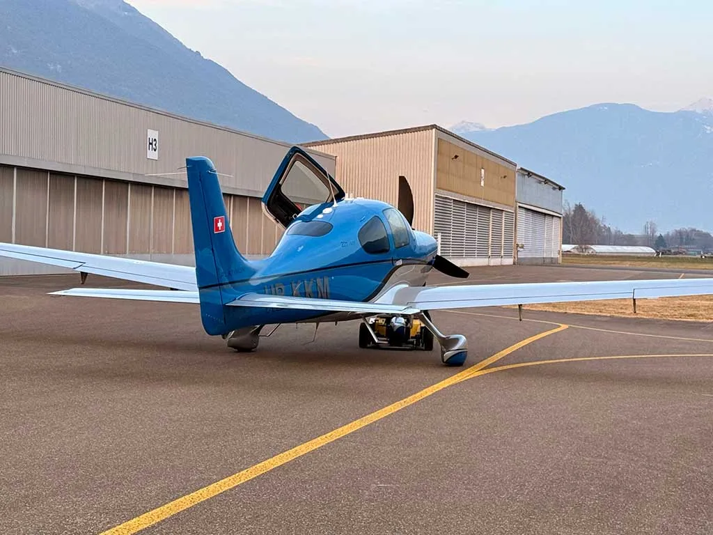 A blue and silver Cirrus SR22T aircraft with its door open, parked on an airfield with hangars and mountains in the background.