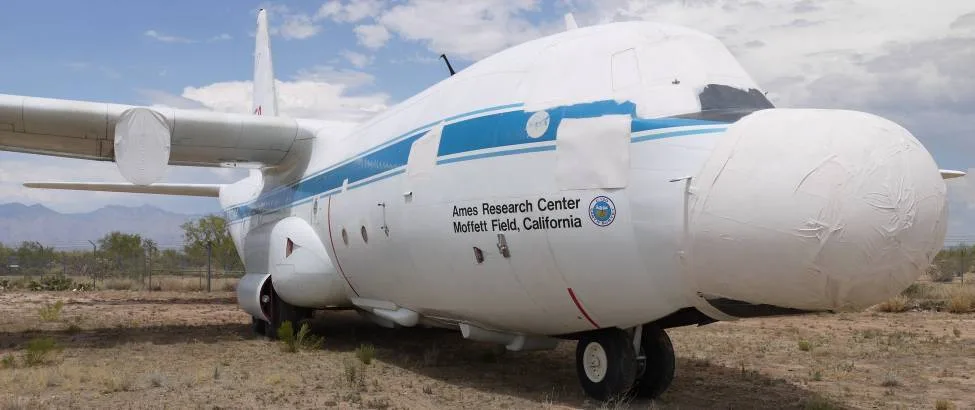 The front-right side of a white NASA C-130 aircraft with covered nose and engine inlets in a desert boneyard.