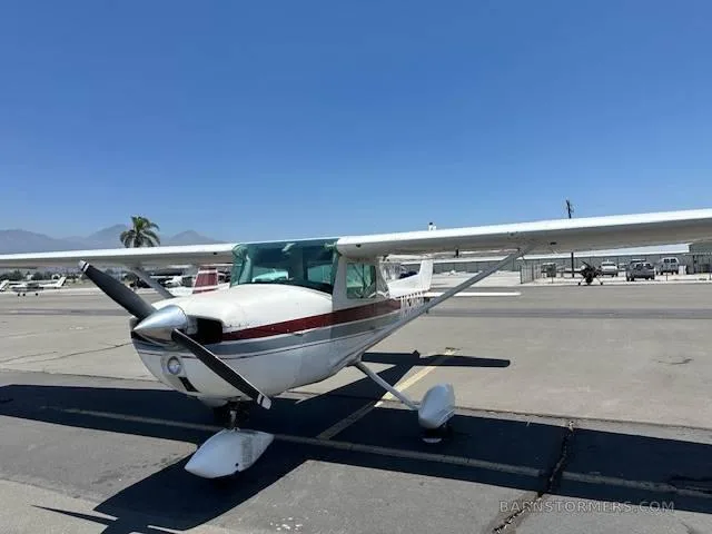 A white Cessna 172 with red and grey stripes is parked on an asphalt ramp, viewed from the front-left.