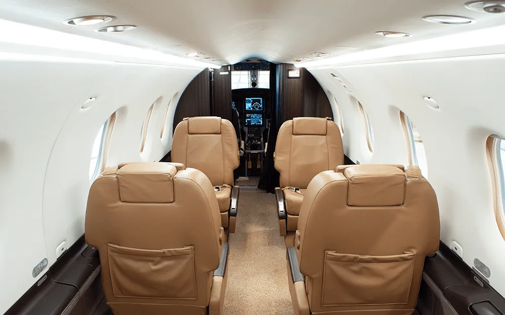 The aircraft cabin interior features two rows of tan leather seats facing forward, with the cockpit visible in the distance.