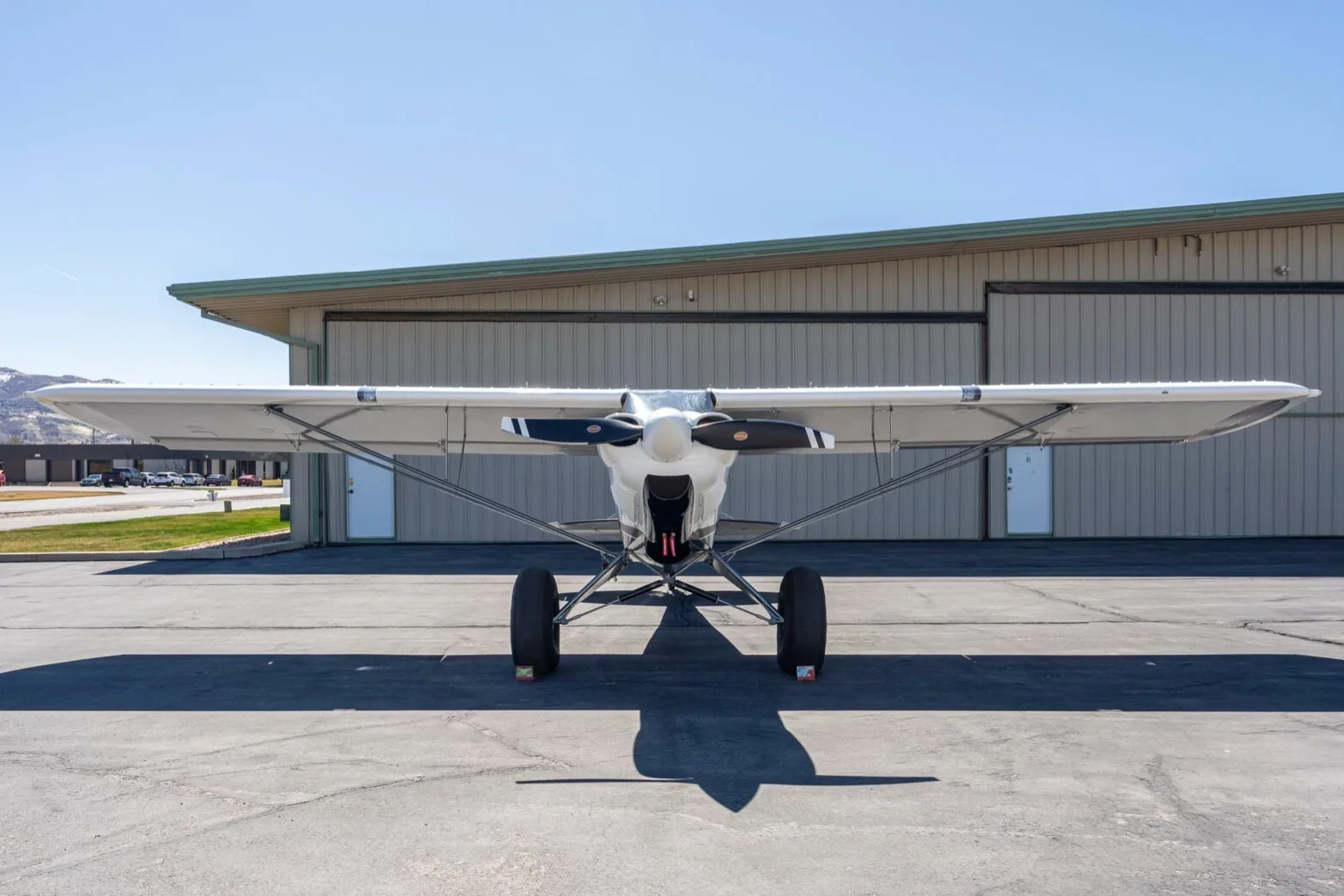 A head-on front view of the white and grey Carbon Cub FX-3 aircraft N25FX parked on an asphalt ramp in front of a hangar.