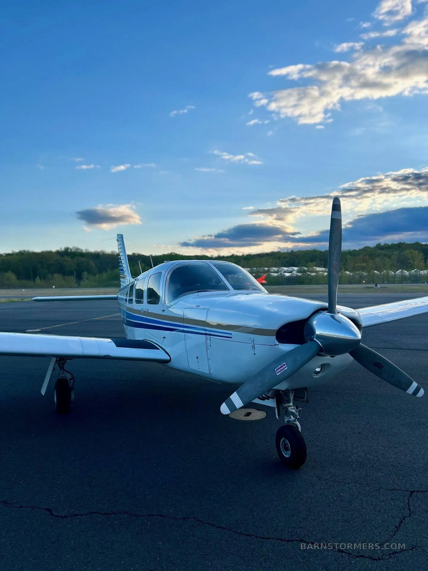 A white single-engine propeller aircraft with blue and gold stripes, viewed from the front-left, on an asphalt tarmac under a blue sky with clouds.