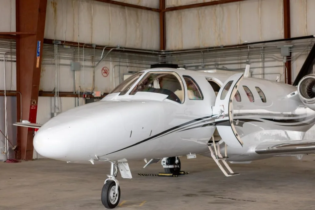 A close-up view of the front of a white Cessna Citation M2 business jet with its cabin door open in a hangar.