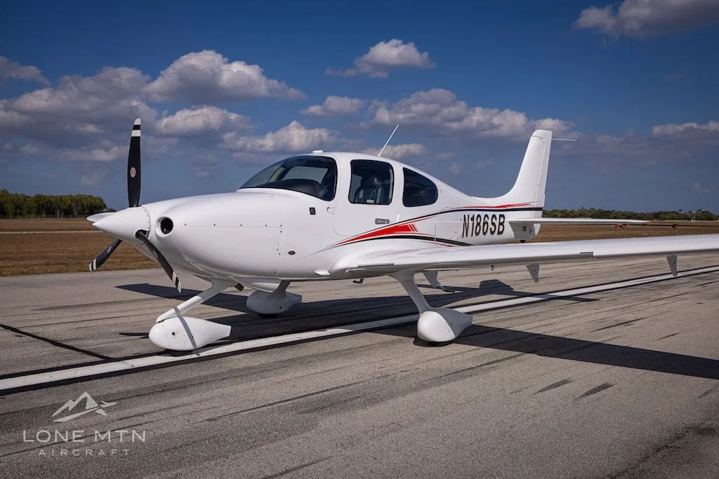 A white single-engine aircraft, N186SB, with red and black accent stripes, parked on an asphalt runway under a blue sky.