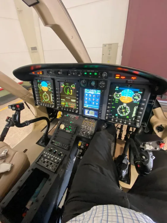 A view of the Bell 429 helicopter cockpit with multiple digital displays and controls, showing a person's legs in the pilot seat.