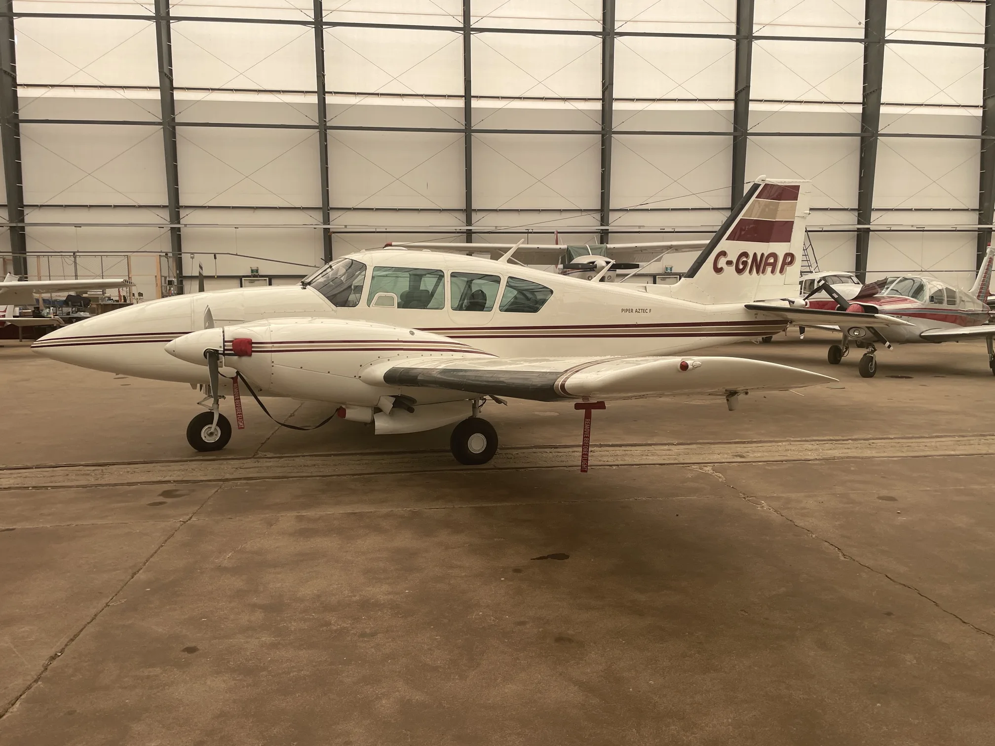 A white Piper Aztec F (C-GNAP) with maroon stripes parked in a hangar.