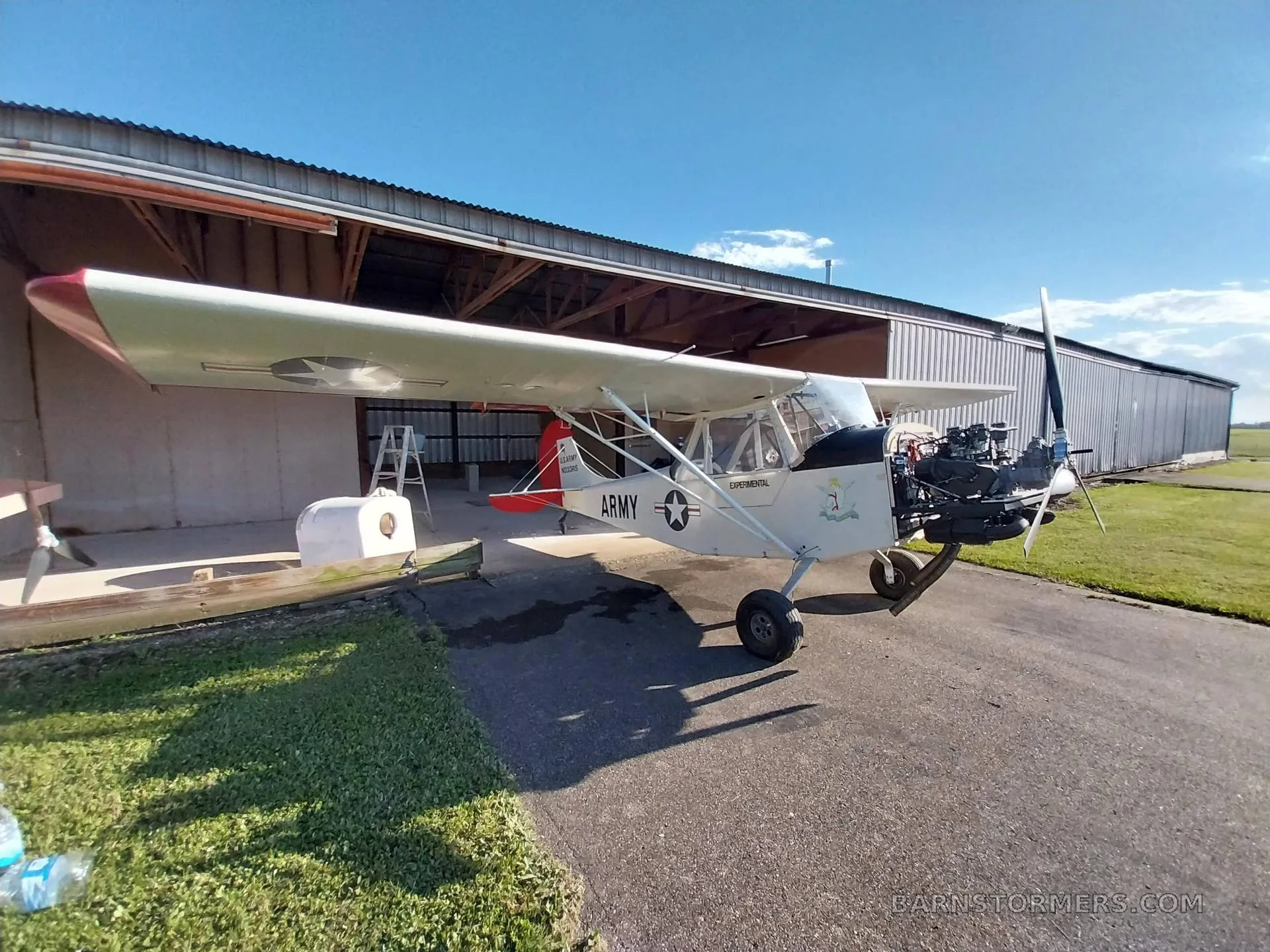 A white experimental aircraft with 'ARMY' markings and an exposed engine sits on an asphalt ramp outside a hangar.
