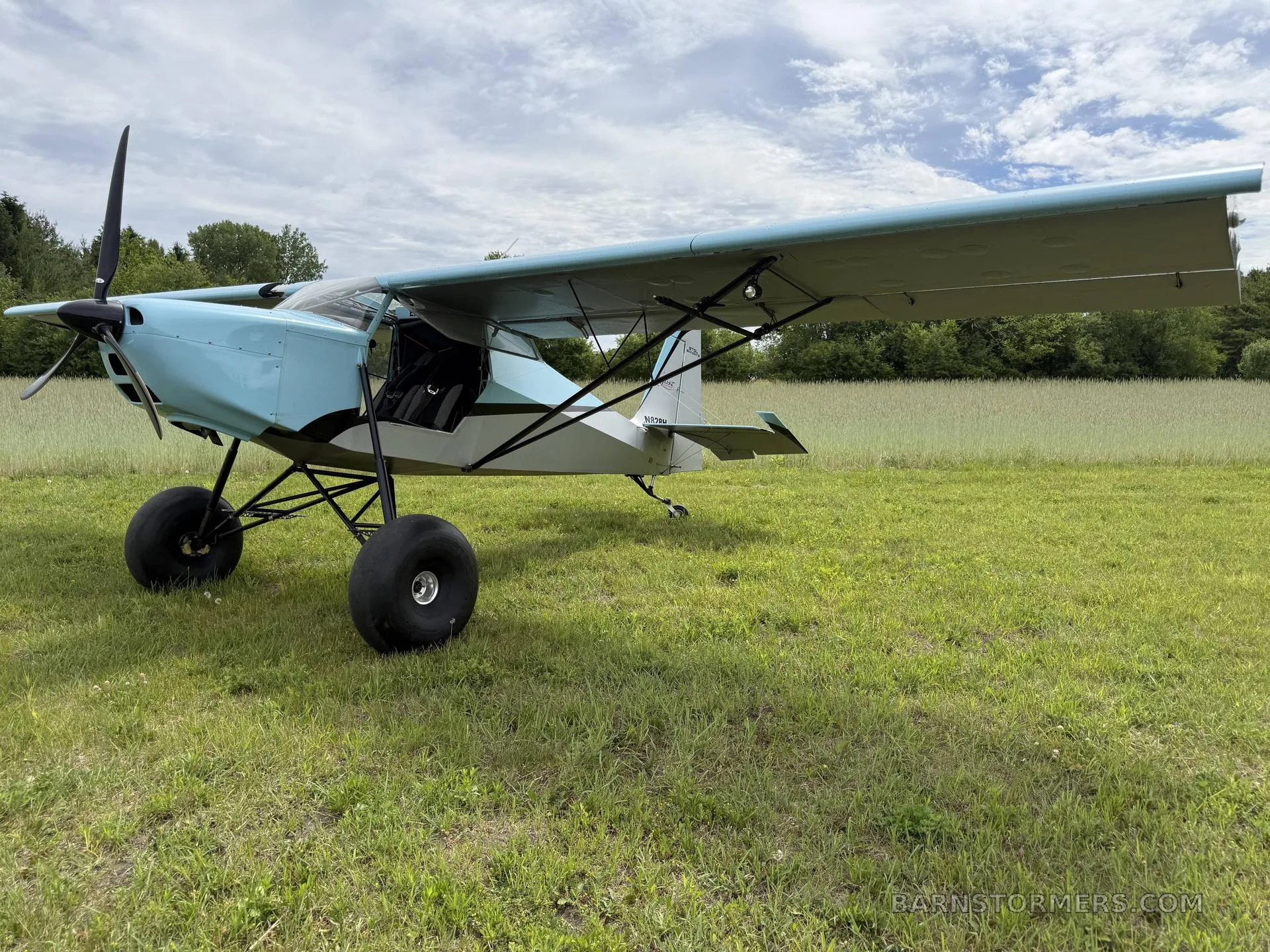 A light blue and grey Just Aircraft SuperSTOL with large tundra tires, parked on a grassy field under a cloudy sky.