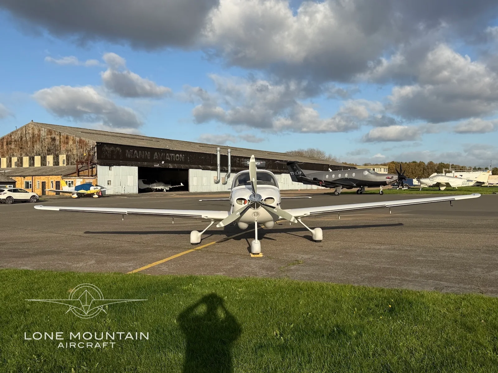 A front view of a white Cirrus SR22 aircraft with a three-bladed propeller, parked on an asphalt apron with hangars in the background.