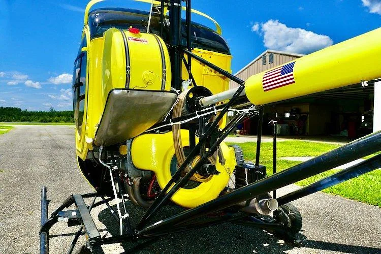 A rear view of the yellow single-engine helicopter, N8872F, showing its tail boom with an American flag decal and the engine compartment.