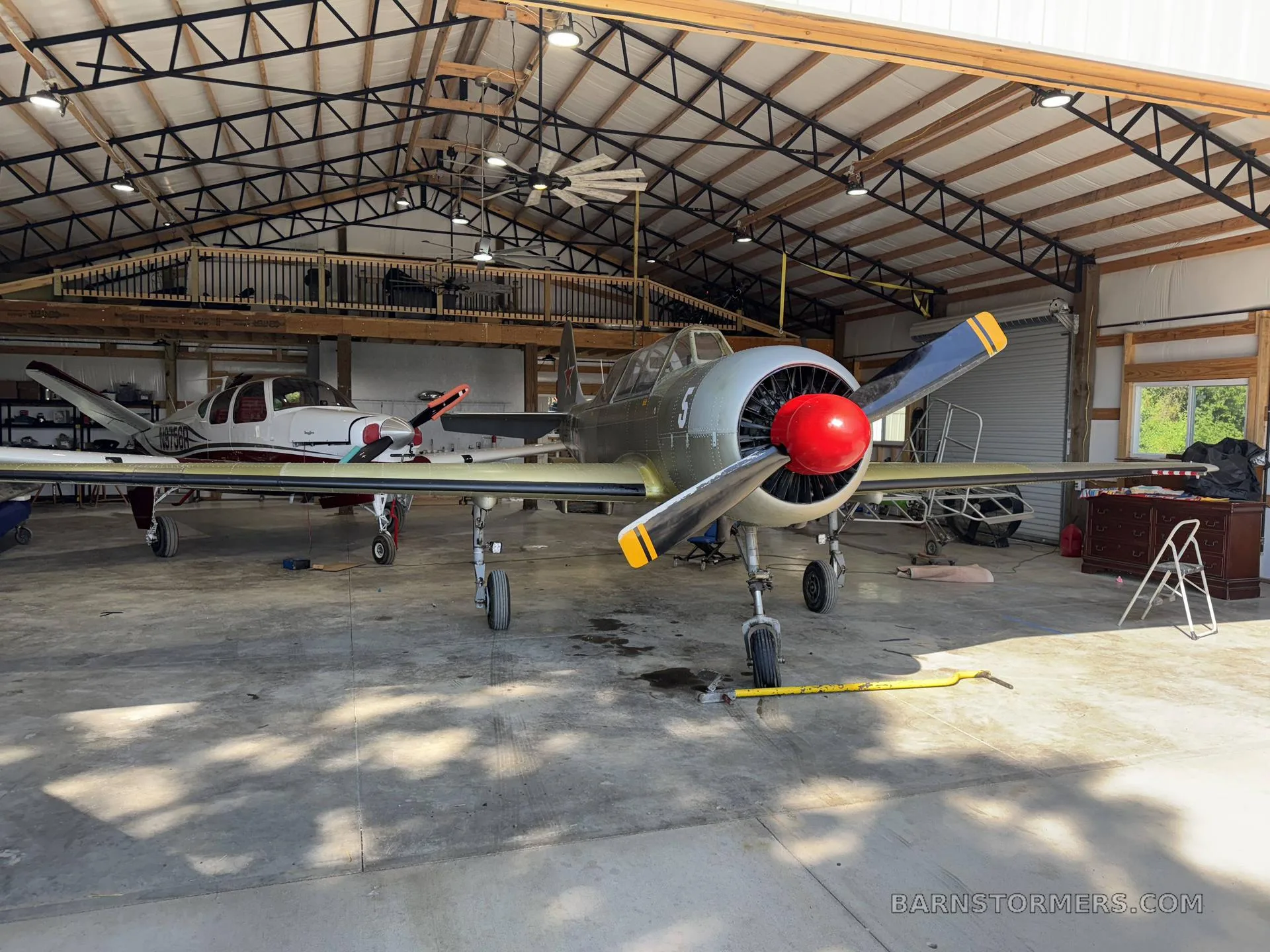 A grey single-engine propeller aircraft with a red spinner and yellow propeller tips, parked in a hangar next to a white and red twin-engine aircraft.