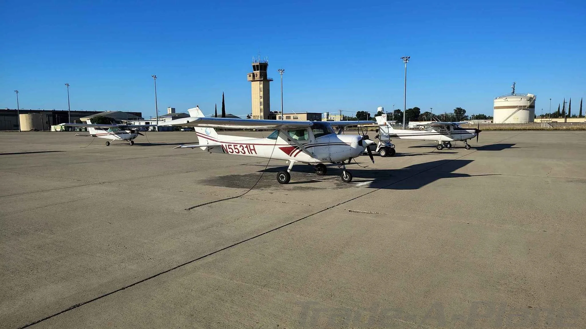 A white Cessna 172 with red stripes, registration N5531H, parked on an airport tarmac with other small aircraft and a control tower in the background.