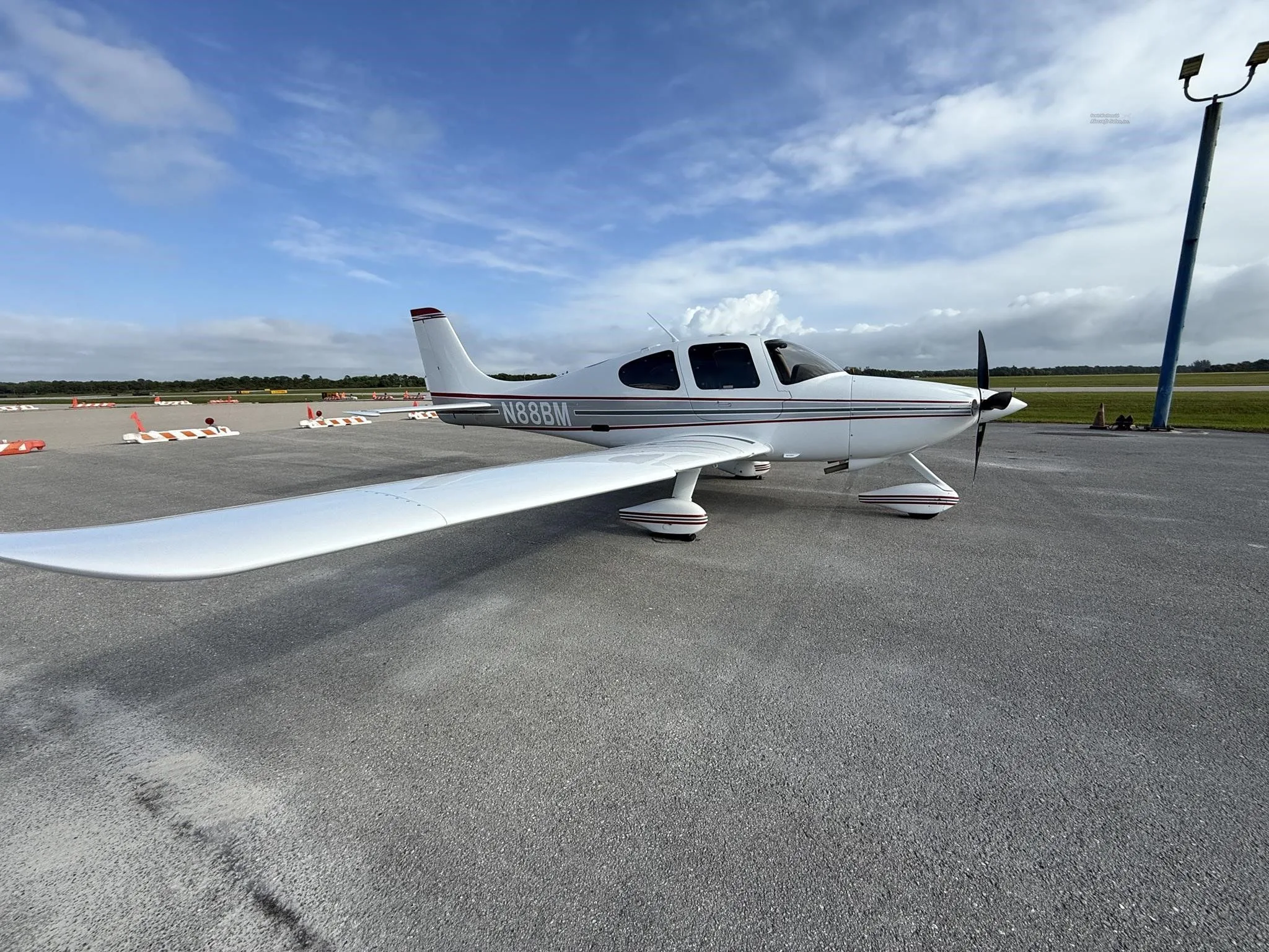 A white Cirrus SR22 aircraft with red and gray stripes, tail number N88BM, parked on an airport tarmac, showing the left side.