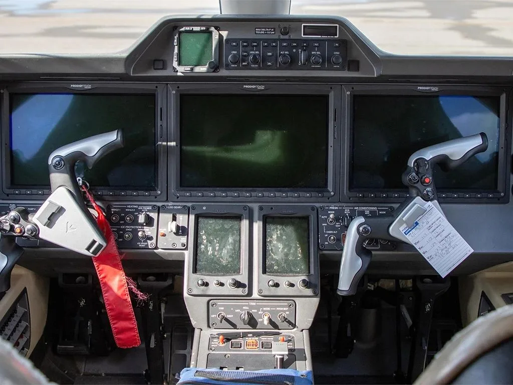 The cockpit of an Embraer Phenom 300, showing multiple display screens, control yokes, and a red tag hanging from one.