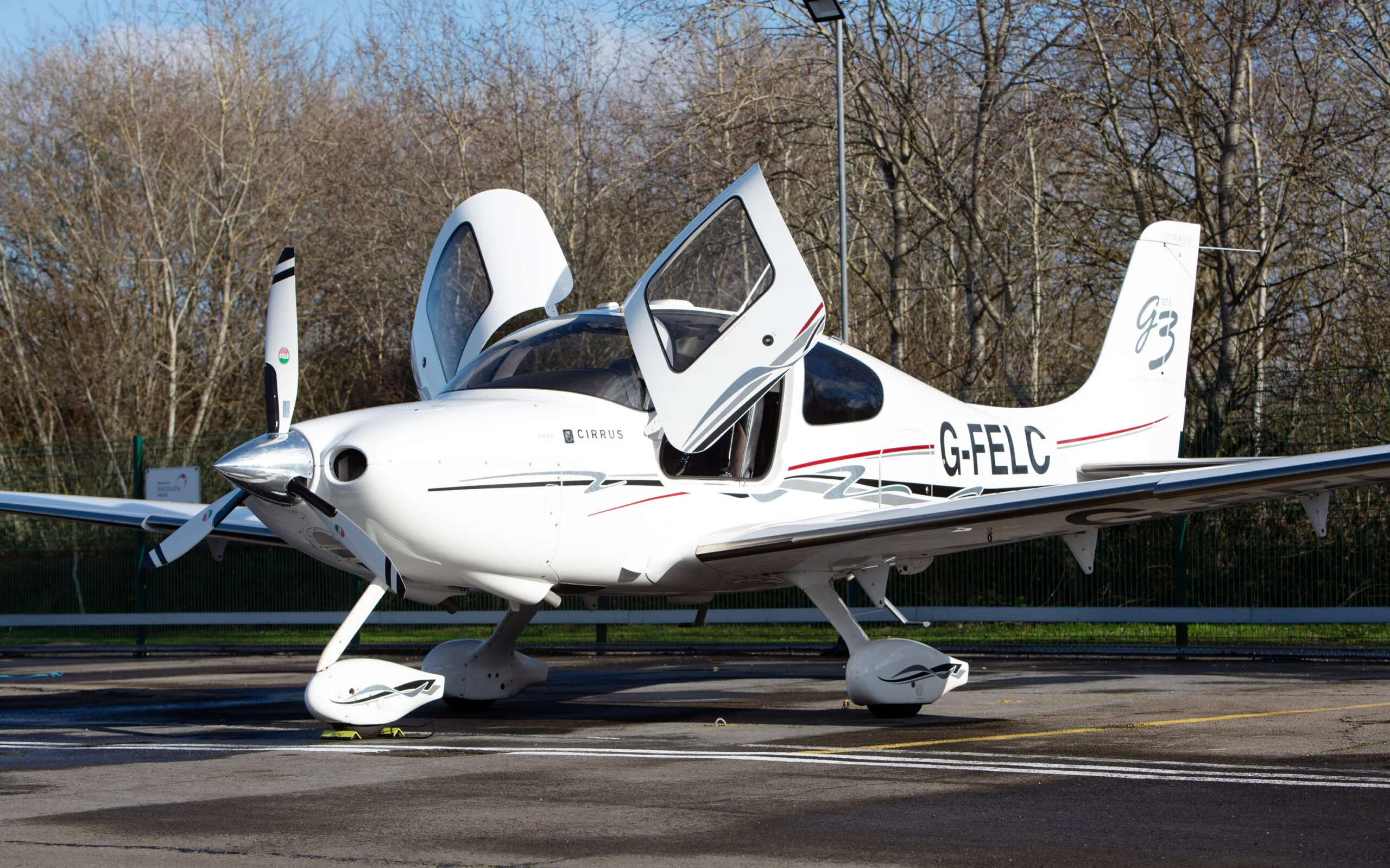 A white Cirrus SR22 G3 aircraft with red and black stripes, registration G-FELC, parked on a tarmac with its gull-wing doors open, viewed from the front-right.