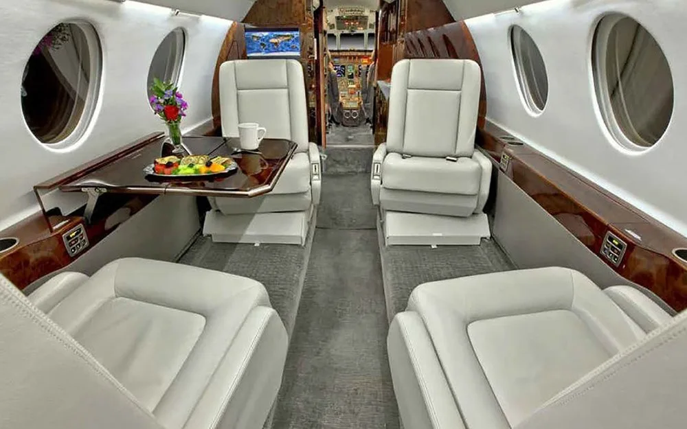 A view of the business jet cabin interior showing light gray seats, a dining table with a meal, and flowers.