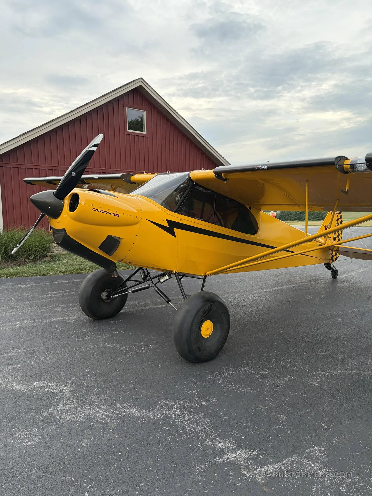 A yellow Carbon Cub aircraft with black stripes and large tires is parked on asphalt in front of a red building.