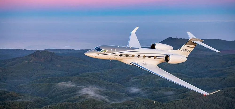 A Gulfstream G500 business jet with a white and grey paint scheme flying over a mountainous, forested landscape at dusk.