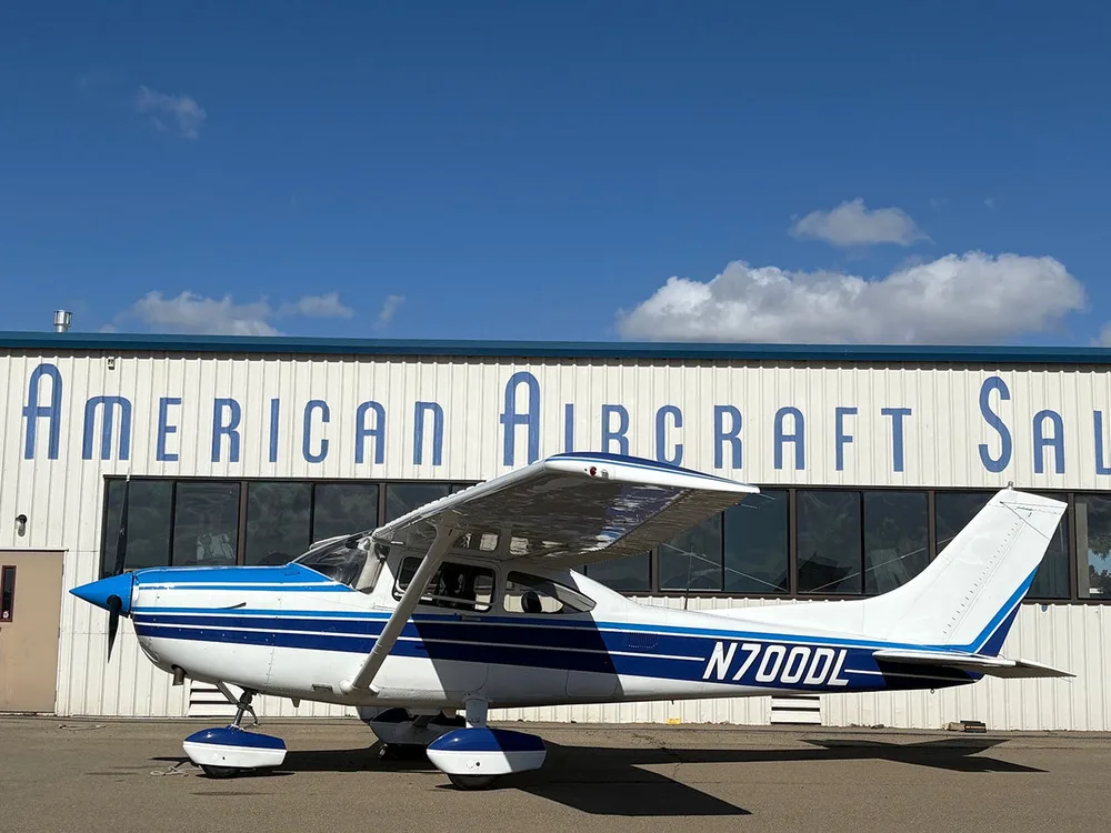 A white and blue Cessna 182 Skylane (N700DL) parked on the tarmac in front of a building.