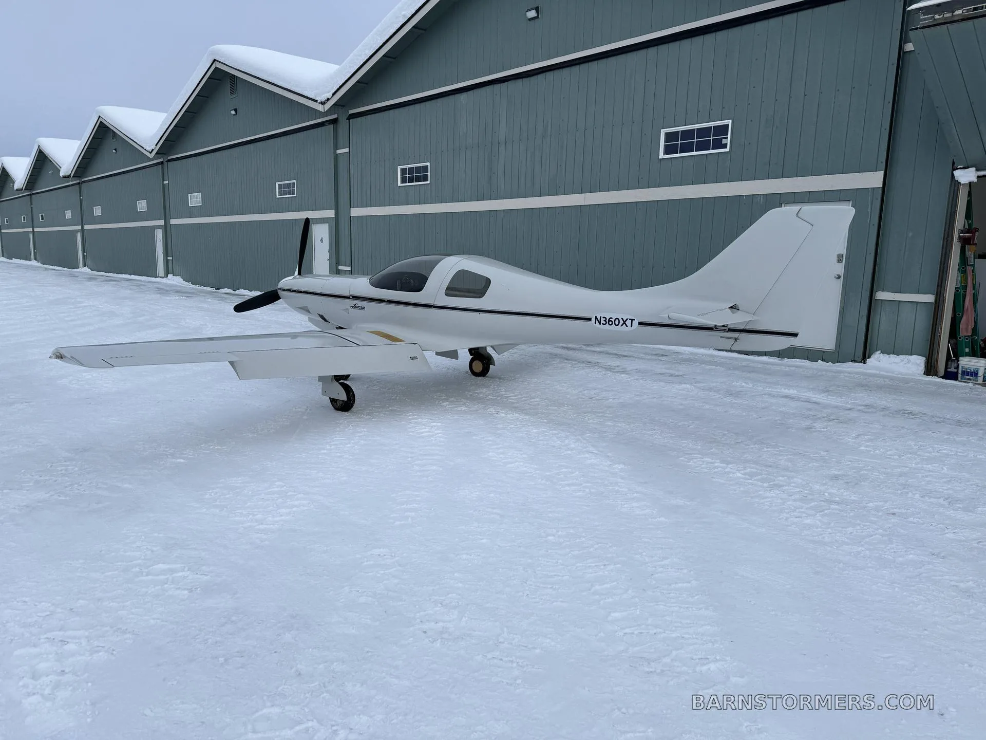 A white Lancair Legacy aircraft (N360XT) with a black stripe is parked on snowy ground in front of a row of blue hangars.