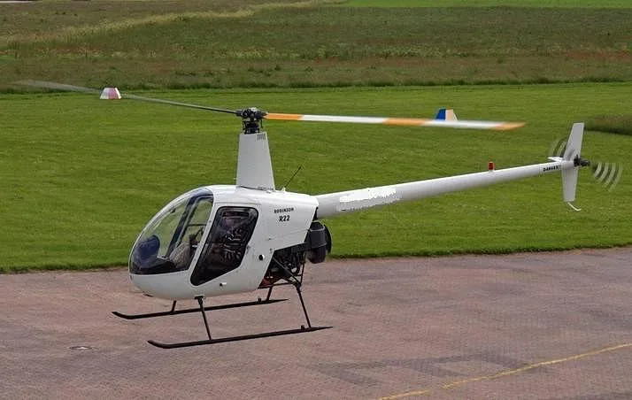 A white Robinson R22 helicopter with spinning main and tail rotors is on a paved surface next to a green field.