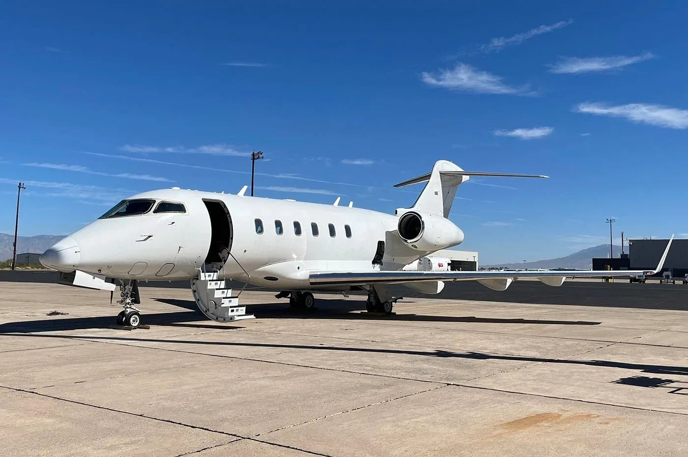 A white business jet, possibly a Bombardier Challenger, parked on an airfield with its main door open and stairs extended, under a clear blue sky.