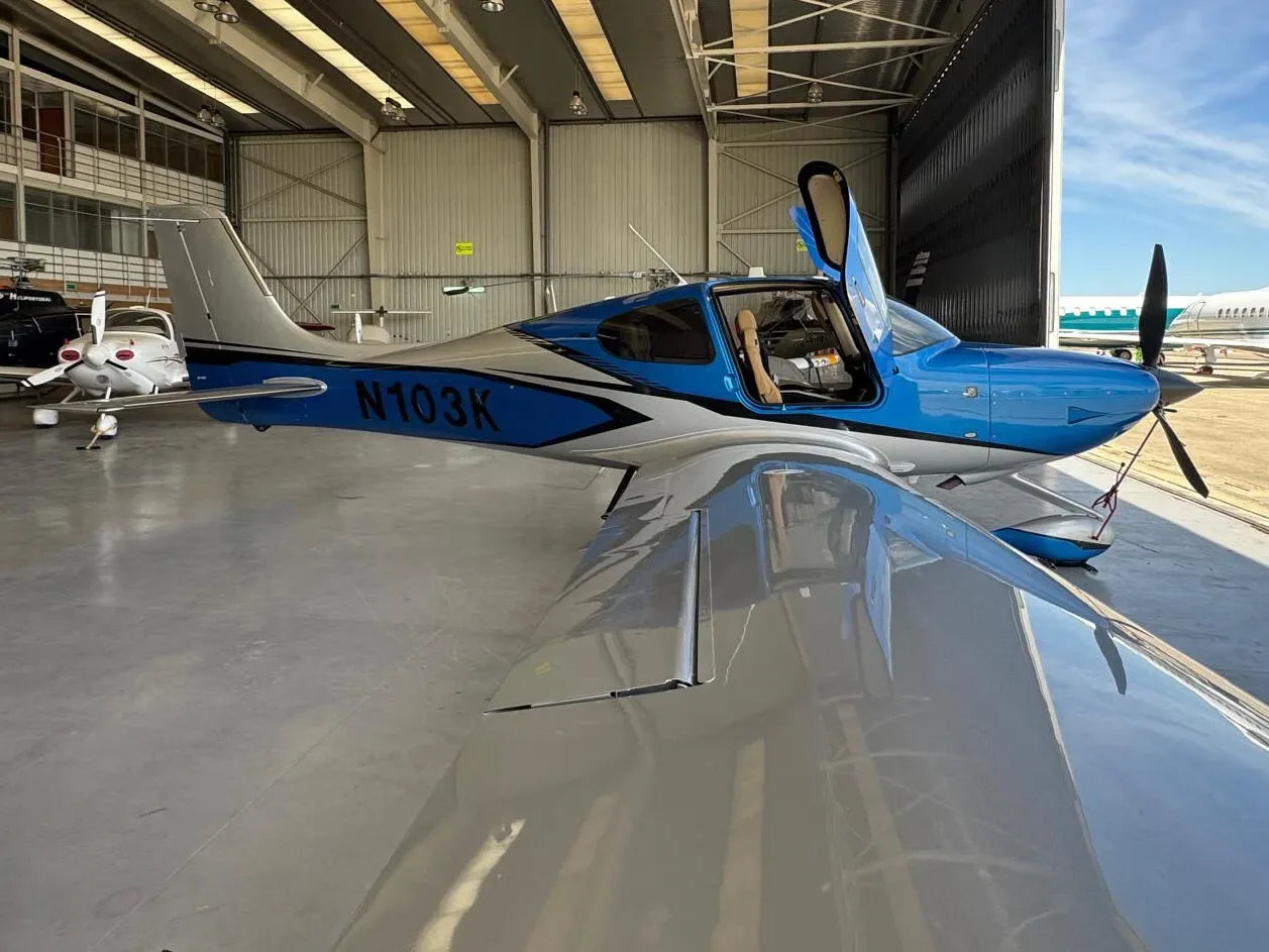 A blue and silver Cirrus SR22 aircraft (N103K) with open doors, viewed from the left side in a hangar.