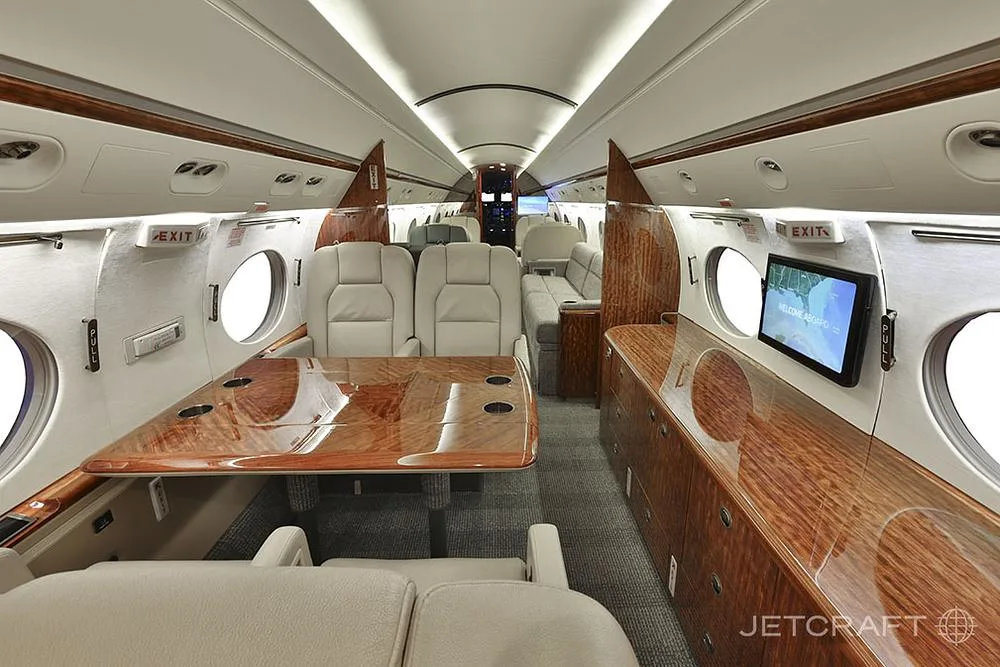 A wide view of the business jet's cabin shows a dining area with a large wooden table and light leather seating.
