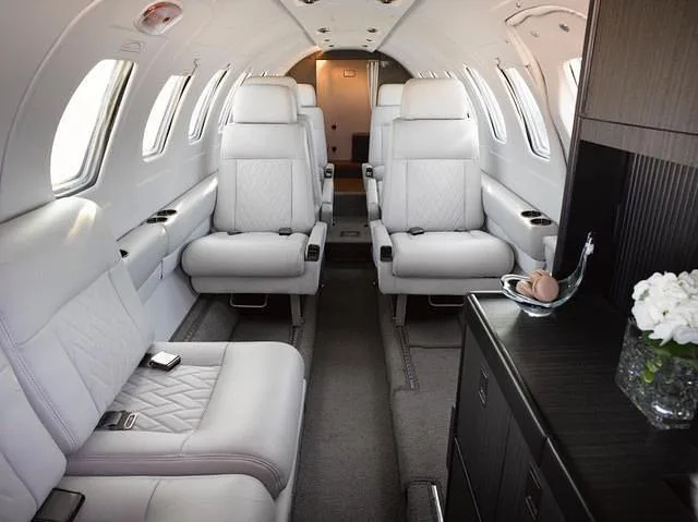 The luxurious white leather interior of a private jet cabin, showing forward-facing seats and a dark wood cabinet.