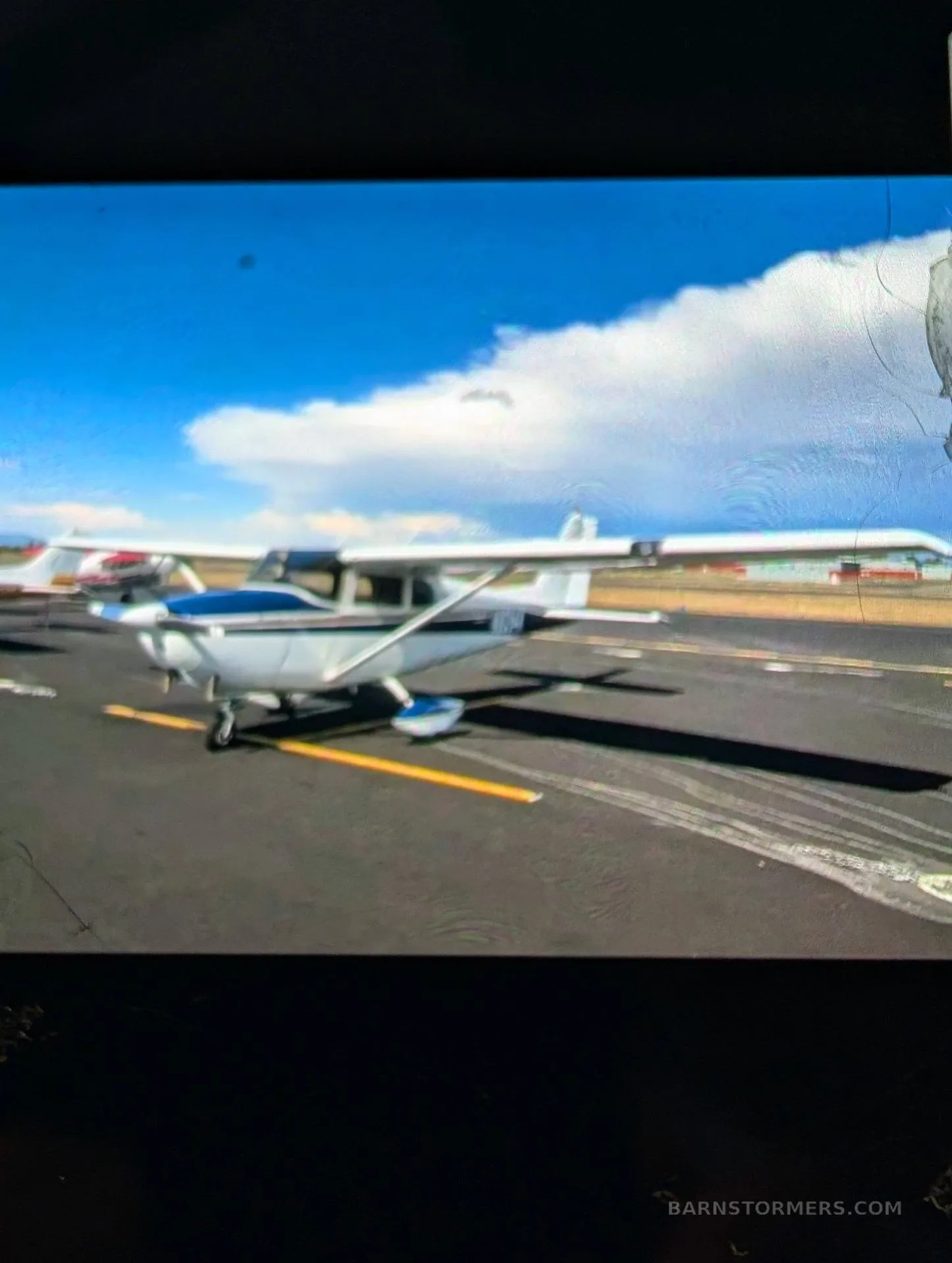 A white and blue single-engine high-wing aircraft, possibly a Cessna, parked on an asphalt ramp under a blue sky with clouds.