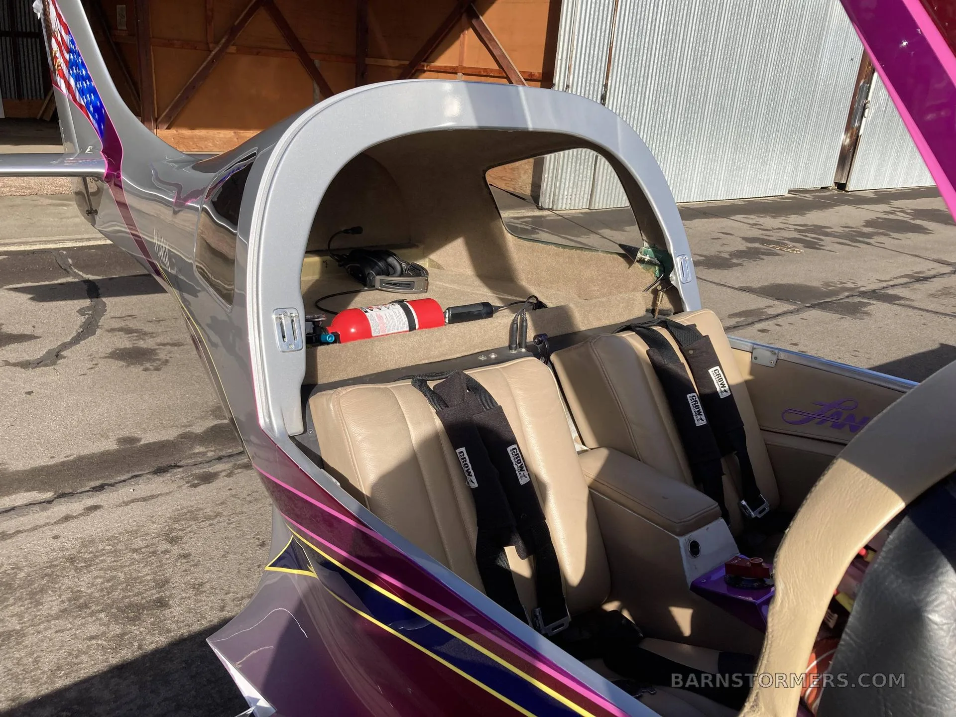 The interior cabin of a Lancair 360 aircraft, showing two tan leather seats with black harnesses and a fire extinguisher in the rear.