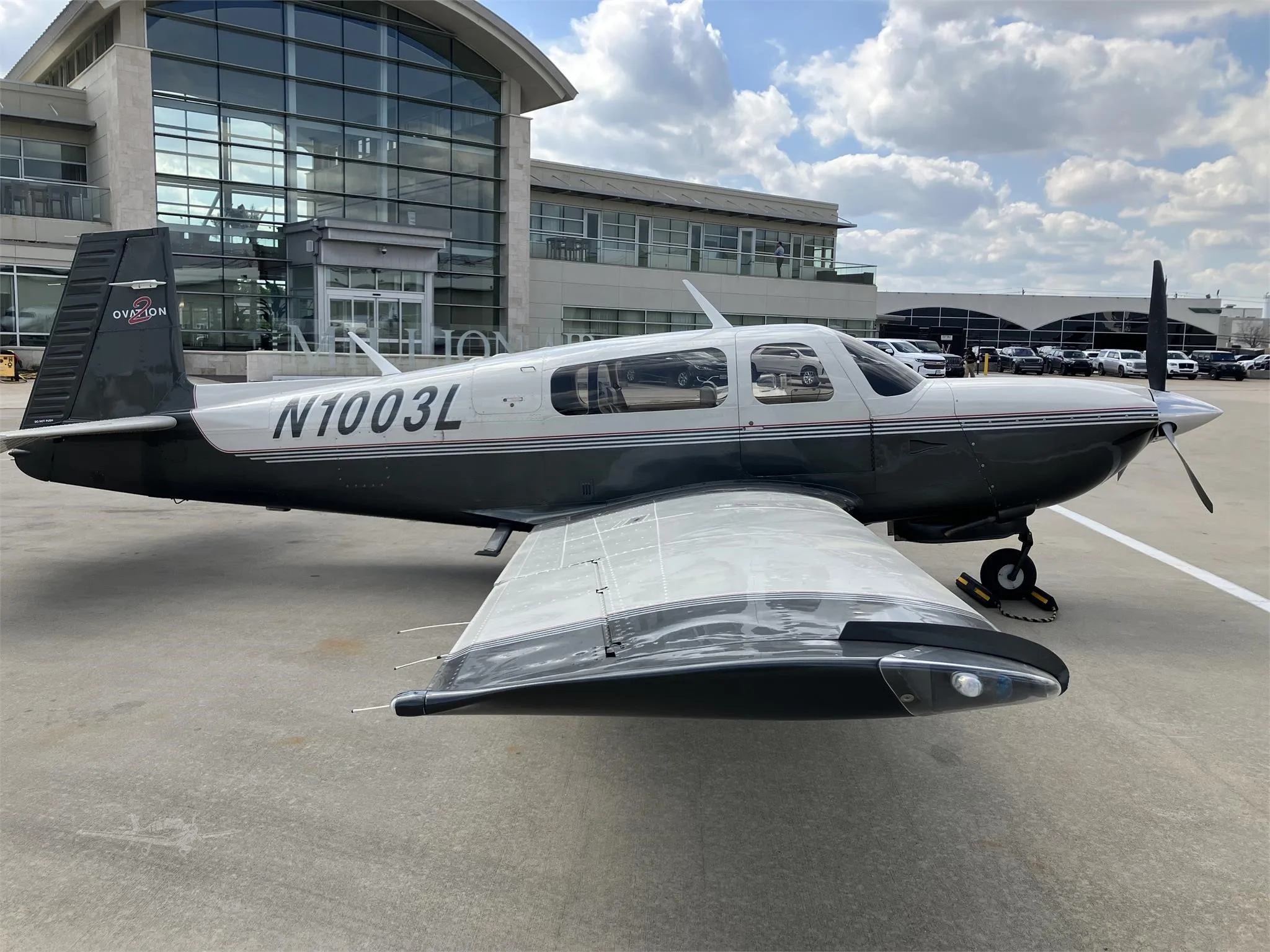 A dark grey and white Mooney M20 Ovation aircraft with registration N1003L is parked on an airport apron in front of a modern building.