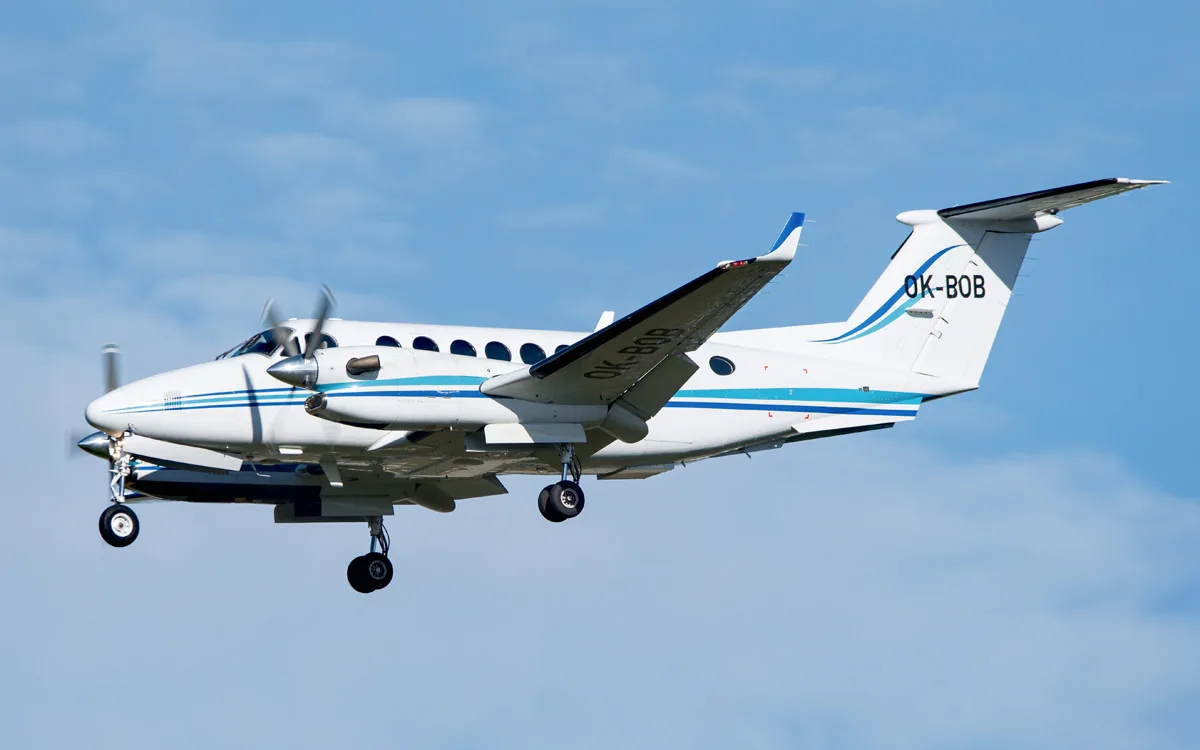 A white twin-turboprop aircraft with blue stripes and registration OK-BOB is shown in flight with landing gear extended against a blue sky.