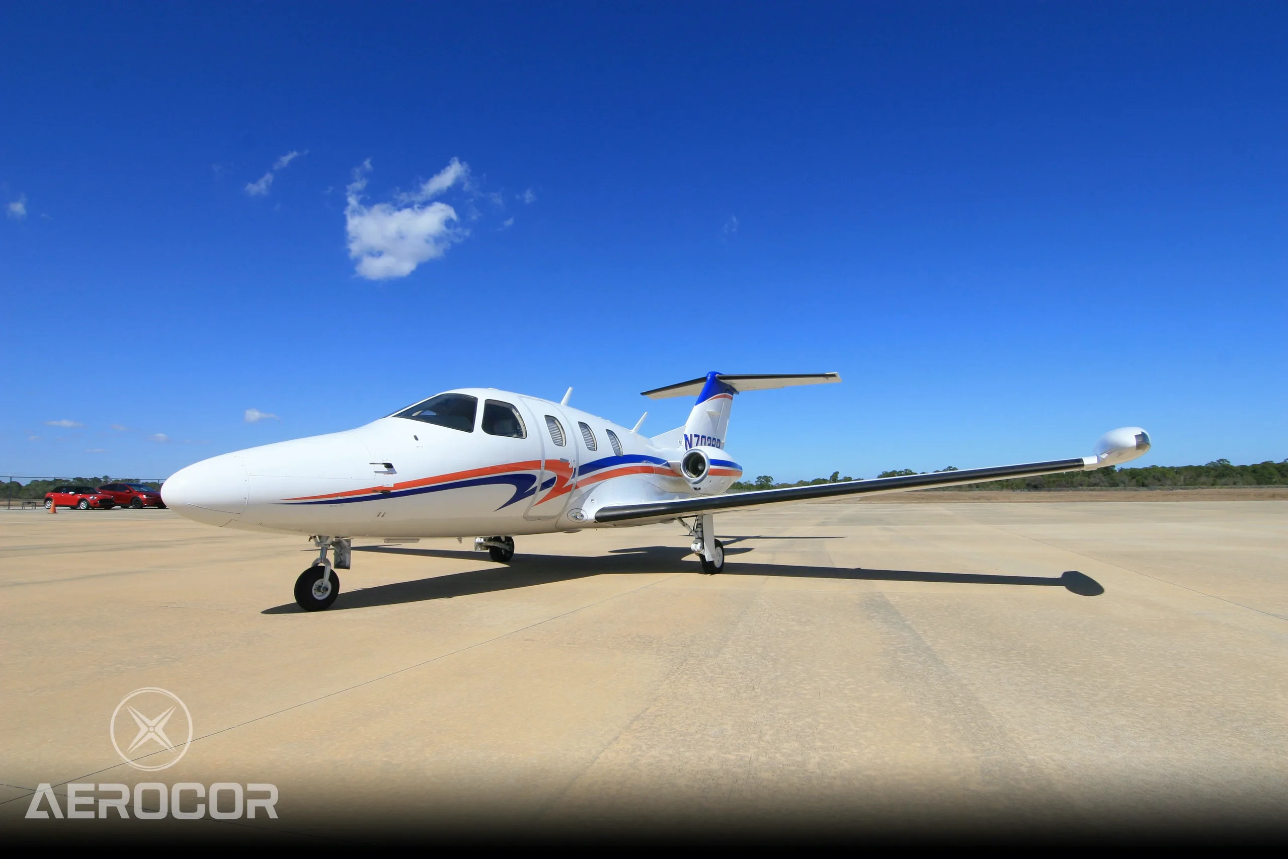 A white Eclipse 500 jet with blue and orange stripes, tail number N703BR, parked on an airfield under a clear blue sky.