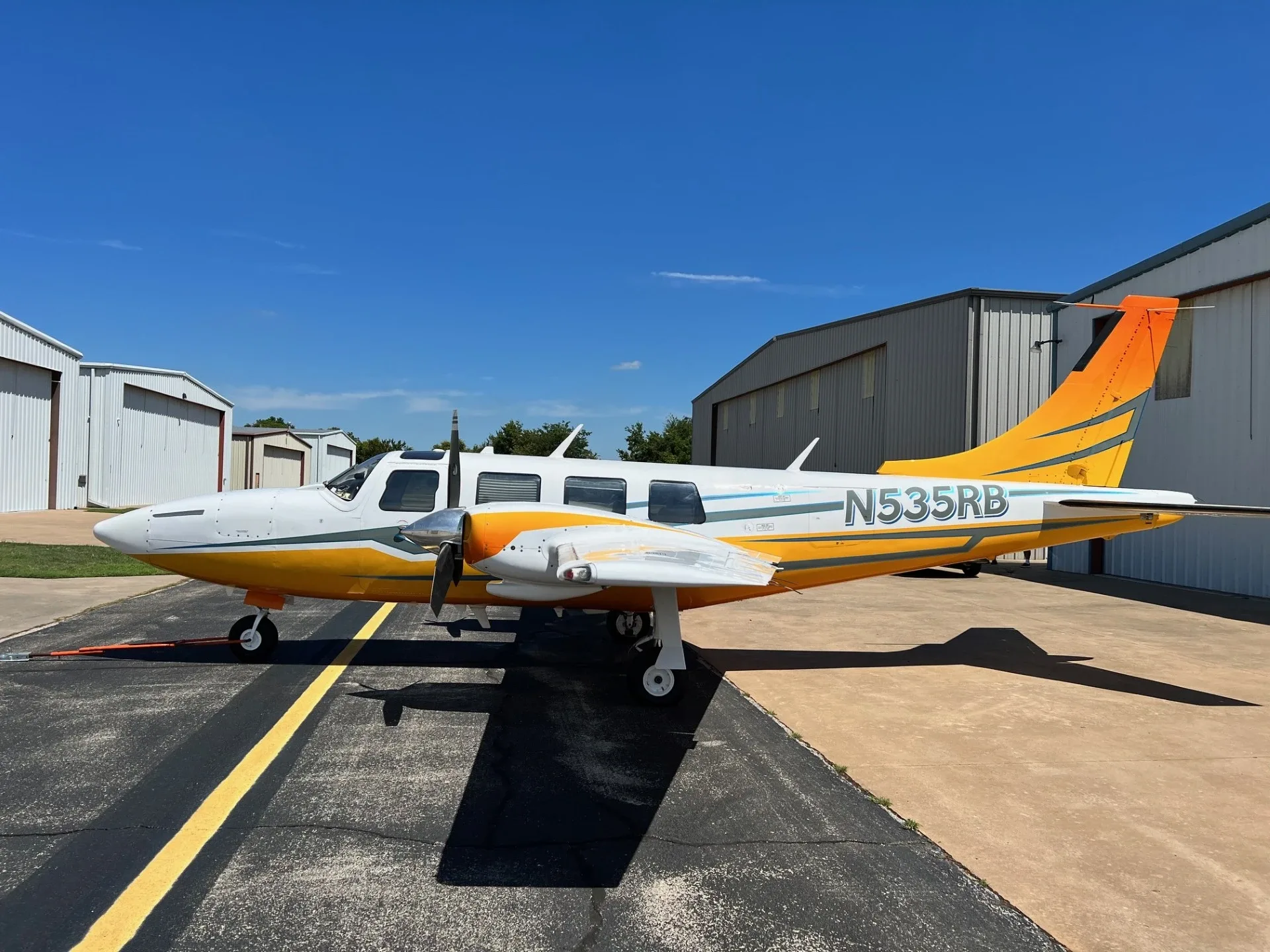 A white and orange twin-engine Piper Navajo aircraft (N535RB) parked on a tarmac next to hangars under a clear blue sky.
