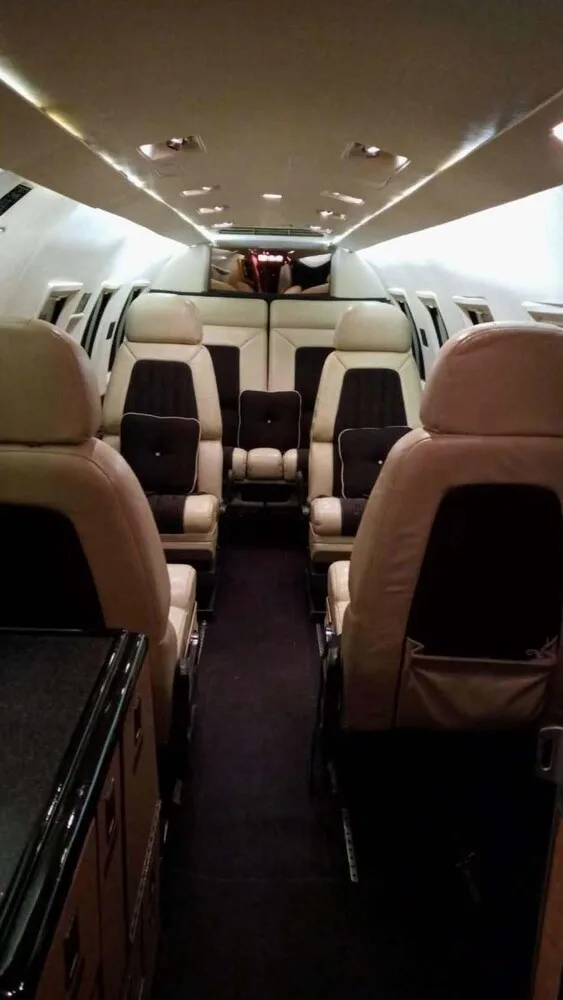 A slightly different angle of the private jet cabin, showing beige and dark brown leather seats and a dark aisle.