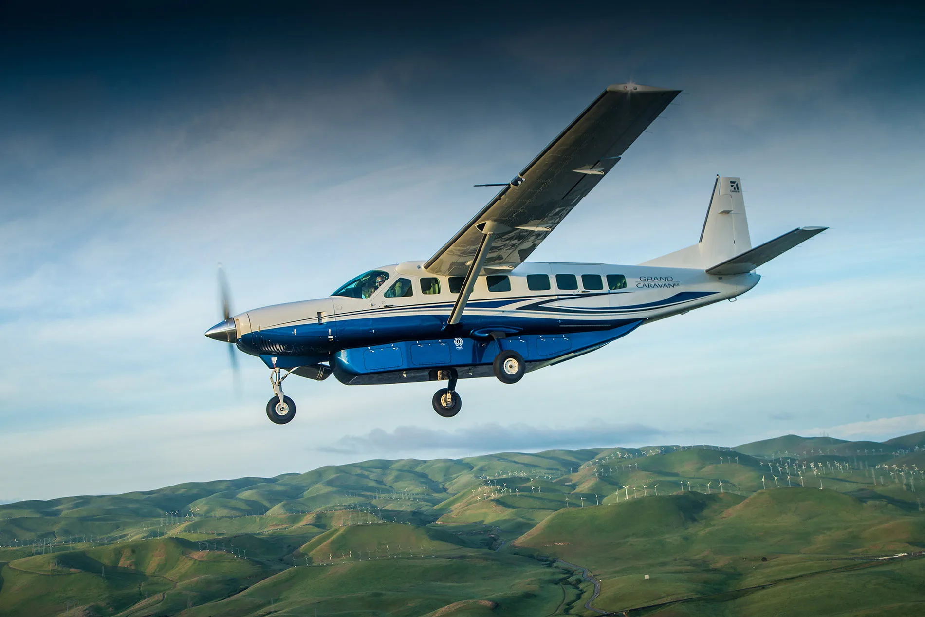 A white and blue Cessna Grand Caravan EX aircraft with landing gear down flies over green rolling hills dotted with wind turbines.