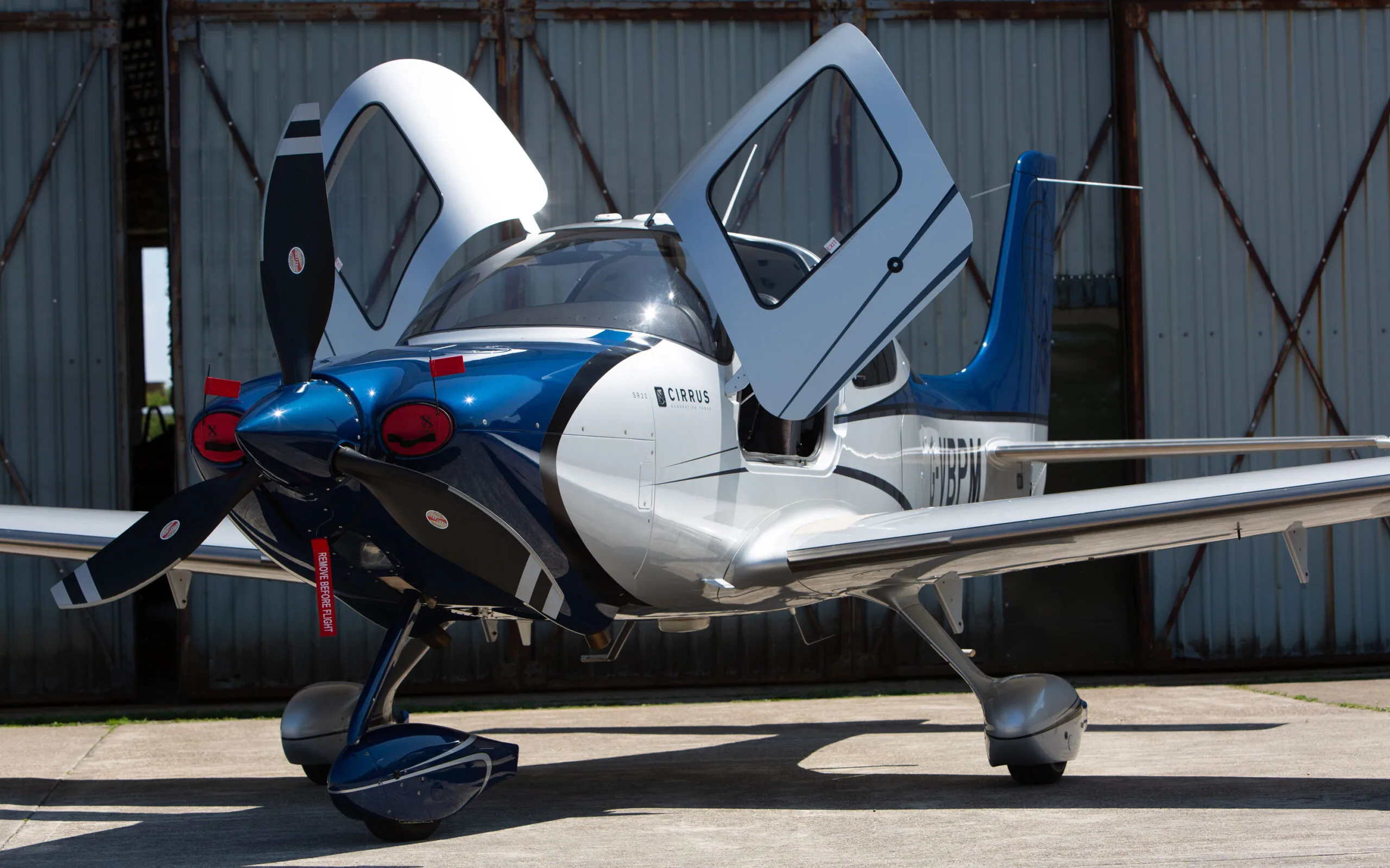 A blue and white Cirrus SR22 aircraft with its gull-wing doors open, parked in front of a hangar.