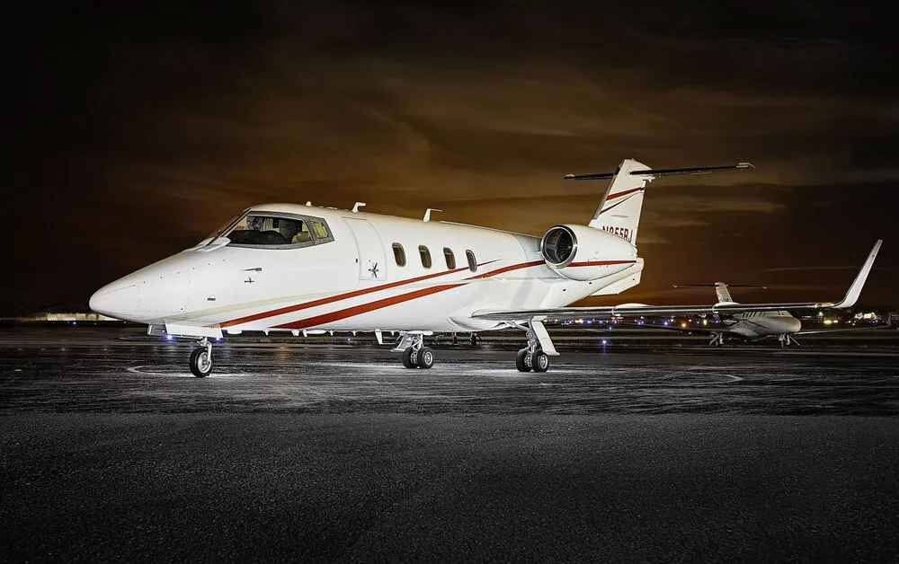 A white Learjet 45 with red and gold stripes, registration N855RJ, parked on a tarmac at night with another aircraft in the background.