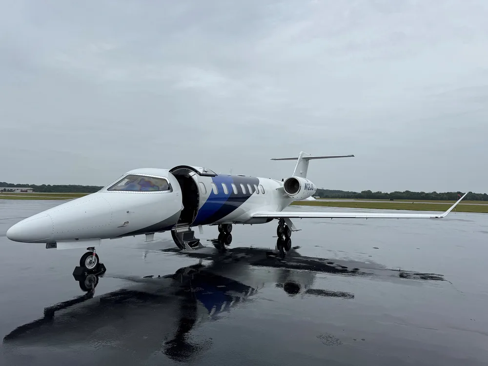 A white business jet with blue and grey accents, registration N12JS, parked on a wet tarmac with its main door open.