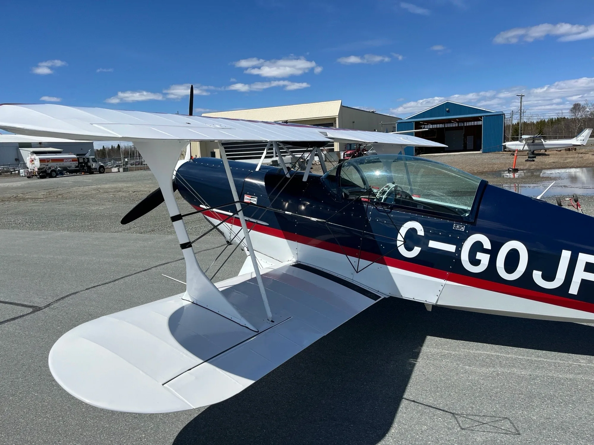 A dark blue biplane with red and white stripes and white wings is parked on an airport tarmac under a blue sky.