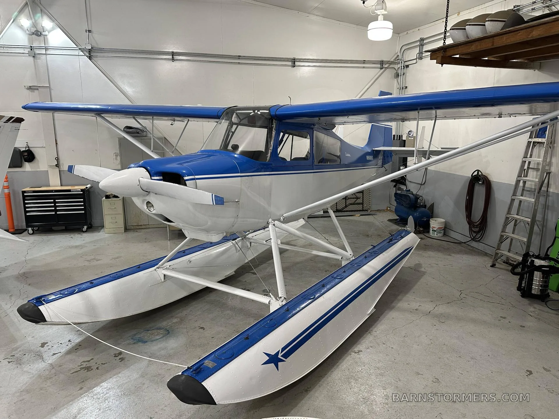 A blue and white Citabria floatplane is parked in a hangar, viewed from the front-left.