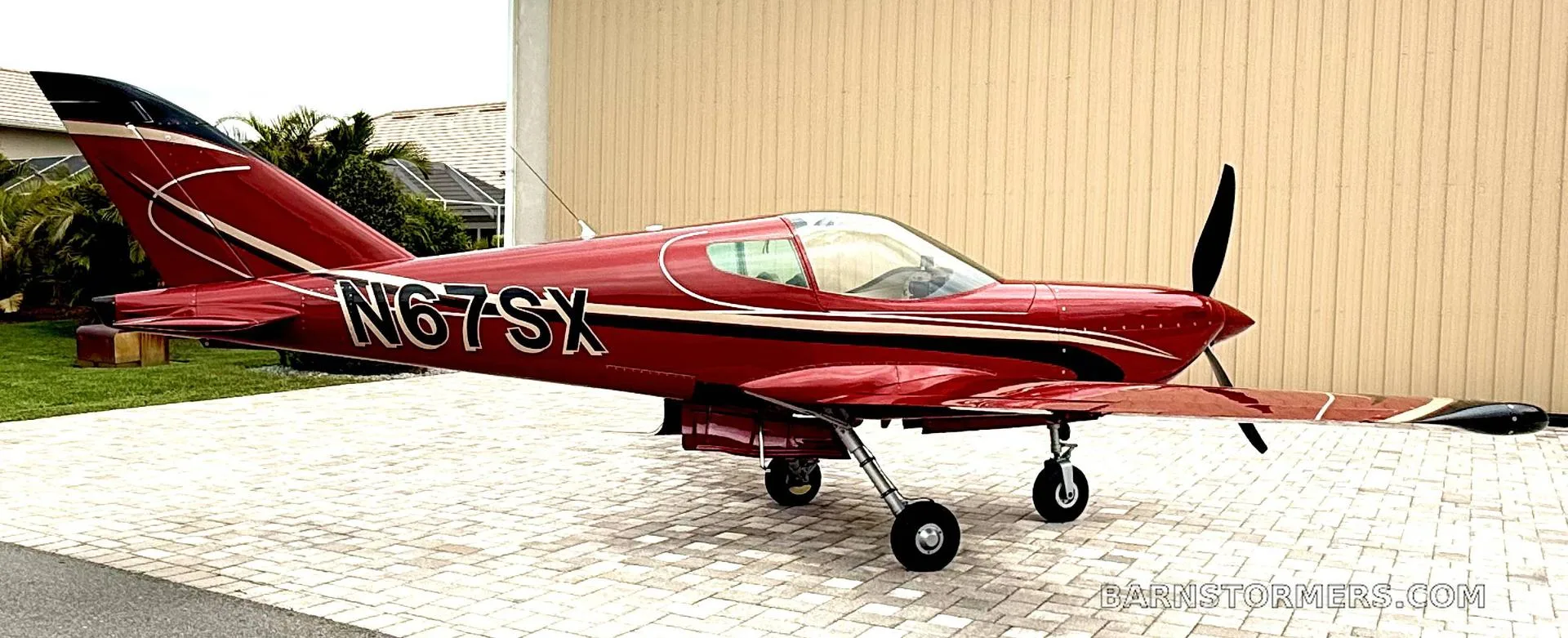 A side view of a red single-engine aircraft with registration N67SX parked on a paved driveway in front of a beige building.