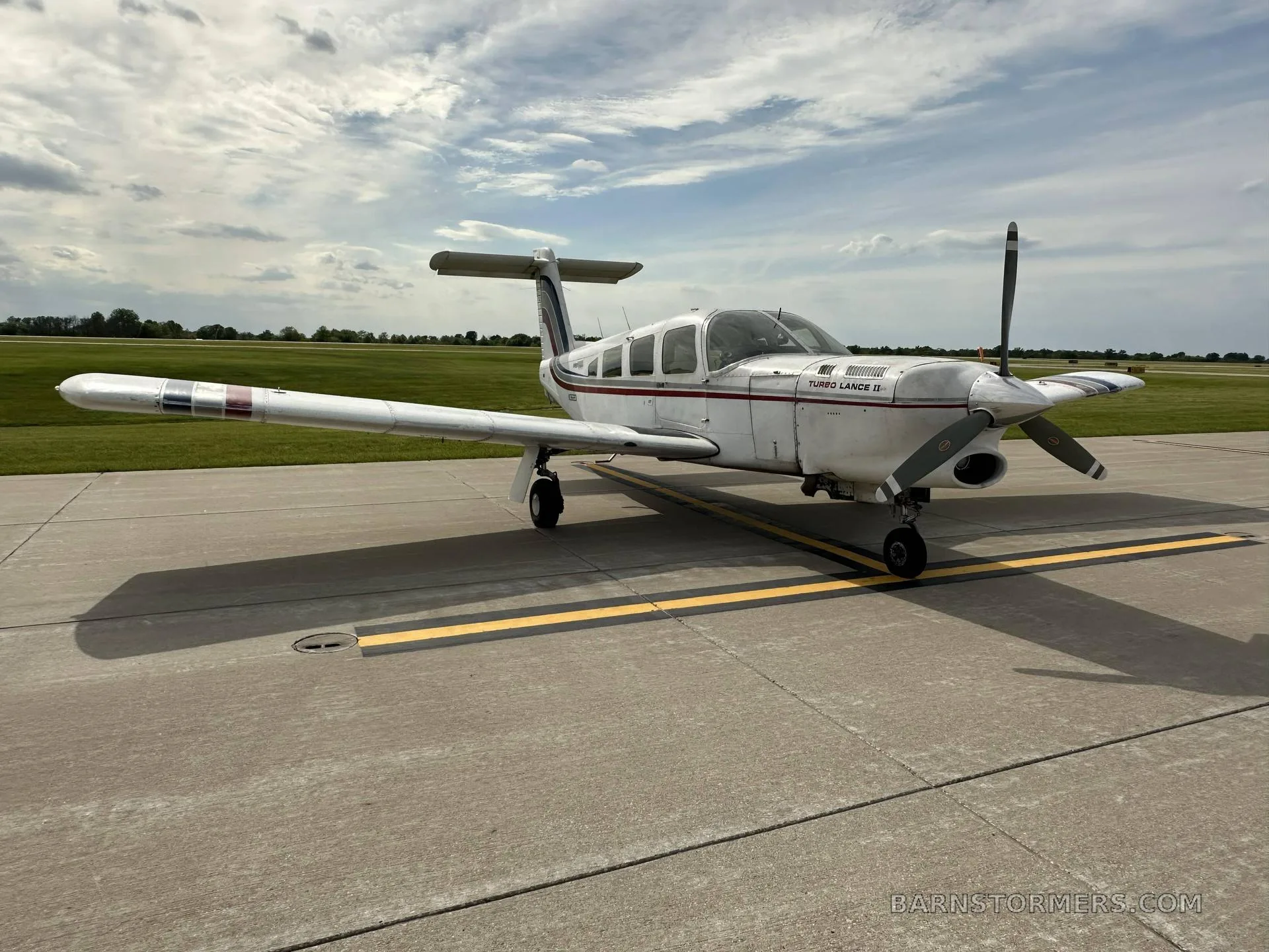 A white Piper Turbo Lance II aircraft with red and blue stripes, parked on an asphalt tarmac under a partly cloudy sky.