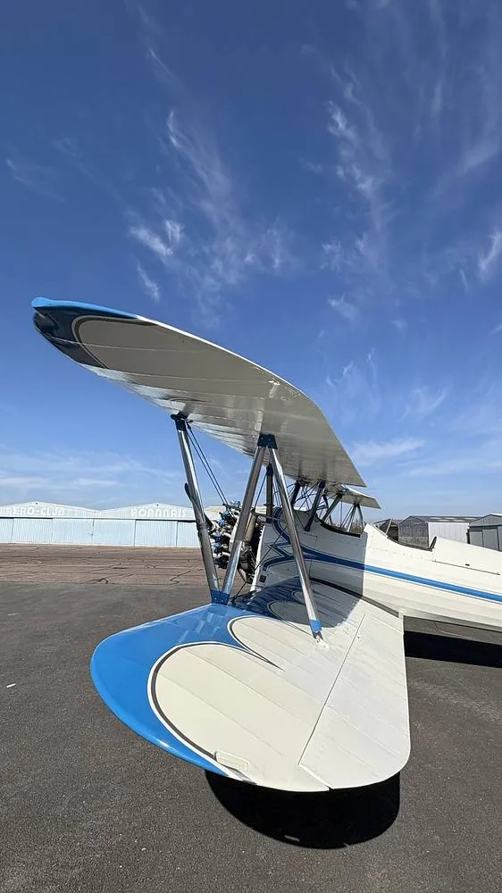 A close-up view of the upper wing and tail of the white and blue biplane, N777CH, parked on an airfield.