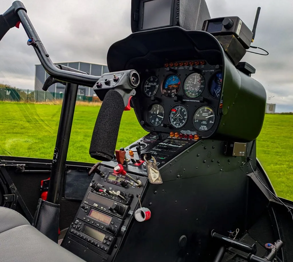 The instrument panel and cyclic control of a helicopter, with a green field visible through the windshield.