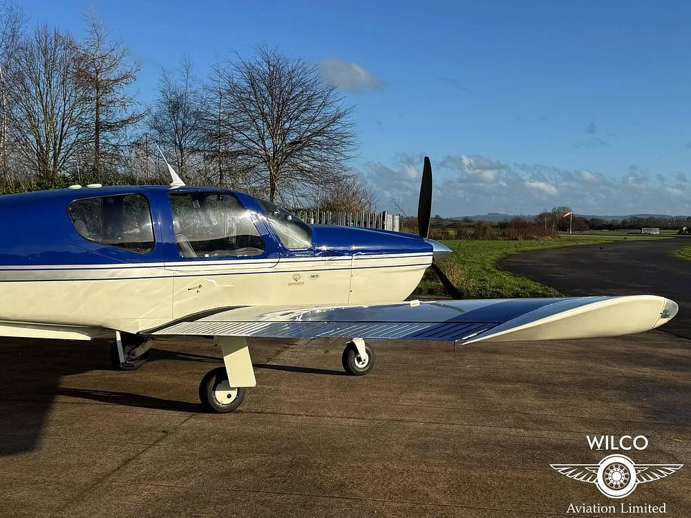 A side view of the blue and white single-engine aircraft, N5ZY, parked on an airfield with a grassy field and distant trees in the background.