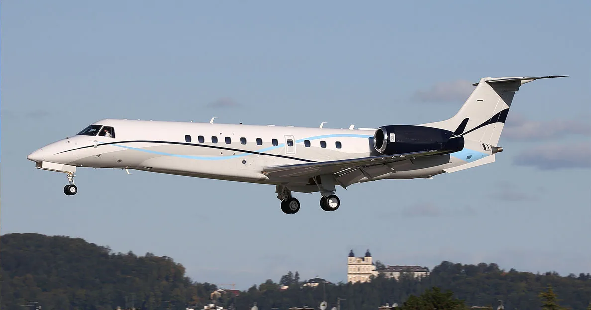 An Embraer Legacy business jet with a white and blue paint scheme, landing gear extended, flying over a landscape with a large building.